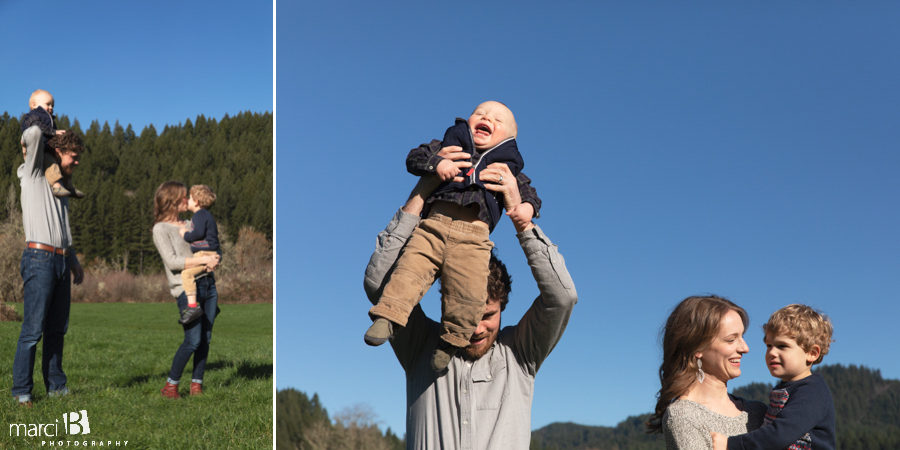 family photographer takes pictures of family with two young boys in Oregon Coast Range - father puts young boy on his shoulders while mother holds four year old son