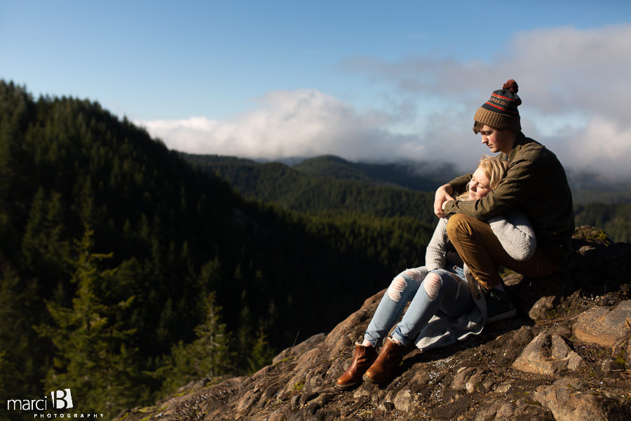 engaged couple sitting on mountain peak above the forest