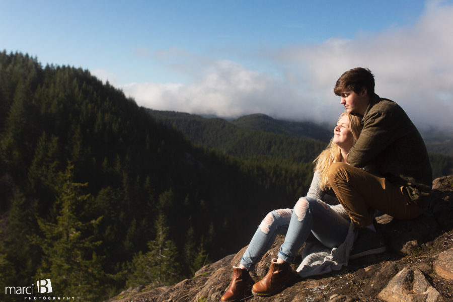 engaged couple sitting on mountain peak above the forest
