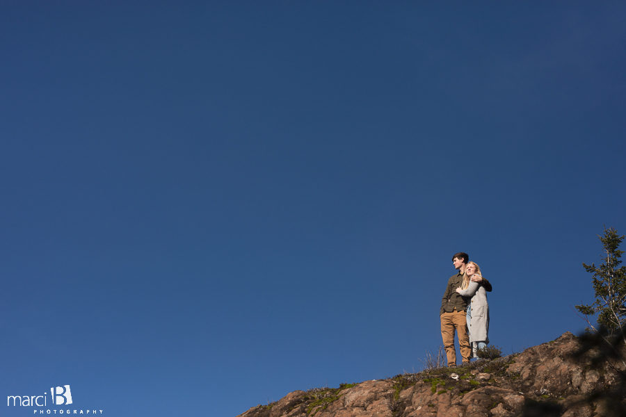 engaged couple standing on rock with blue sky in background
